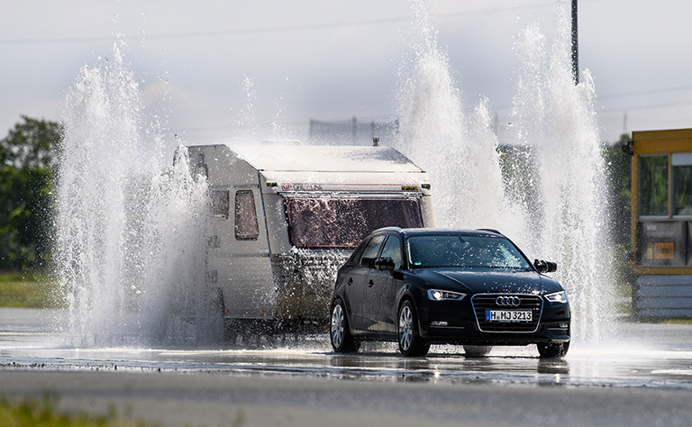 На фото: автомобиль с прицепом проезжает через фонтан, бьющий из-под колес, во время обучения правилам безопасности вождения.
