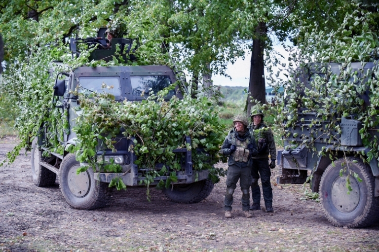 На фото: украинские военные стоят возле военной техники, спрятанной под ветвями деревьев.